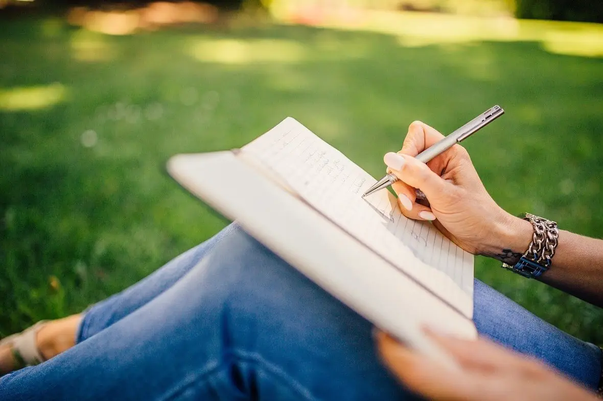 Close-up of a person's hands writing in a notebook while sitting on the grass