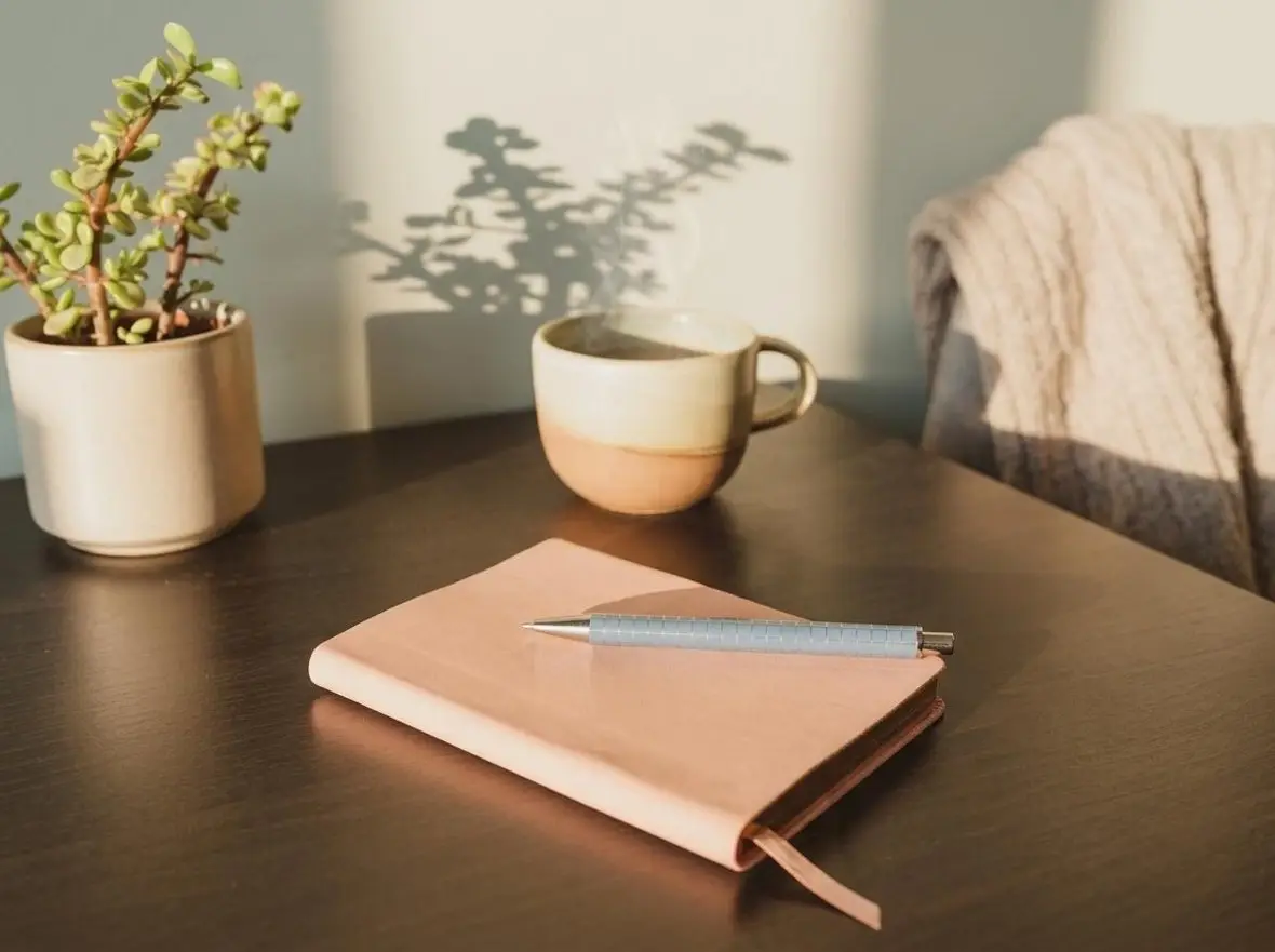 A notebook, pen and mugs on a dark wooden table beside a plant