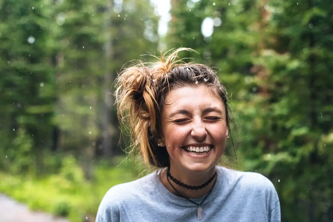 A person laughing outdoors in front of green trees