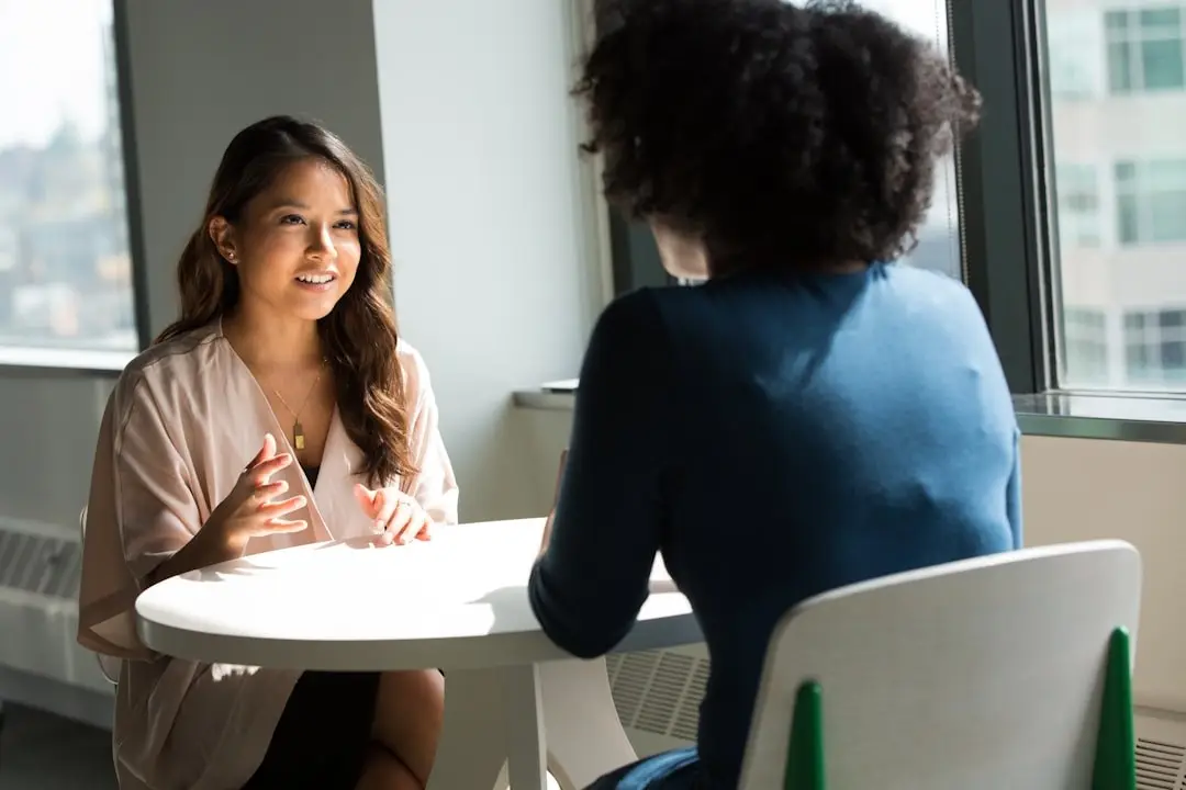 Two people talking across a small table beside a window