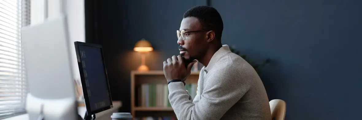 A man in glasses sitting at a desk, chin in hand, looking thoughtfully at a computer screen
